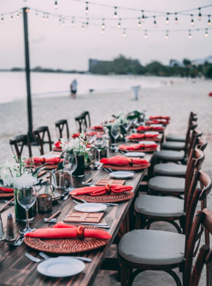 Velika lesena miza in stoli na plaži, obložena z meniji, dekracijo, rdečimi prtički, krožniki in kozarci, pripravljena za večerjo.